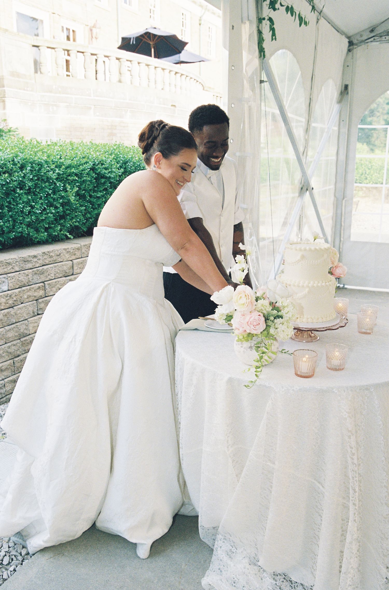 Bride and groom cutting white wedding cake together under tent at outdoor reception with floral arrangement.