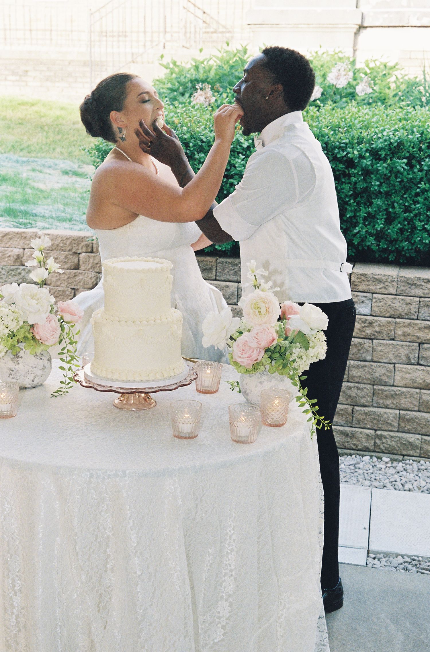 Bride and groom sharing a joyful cake cutting moment at their elegant outdoor wedding reception with white floral arrangements.
