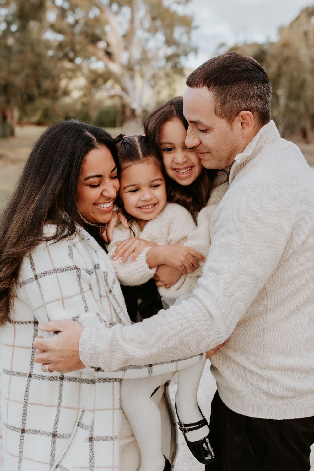 Young parents dressed in warm knits hug their little girls in a field at golden hour for family photos with professional photographer.