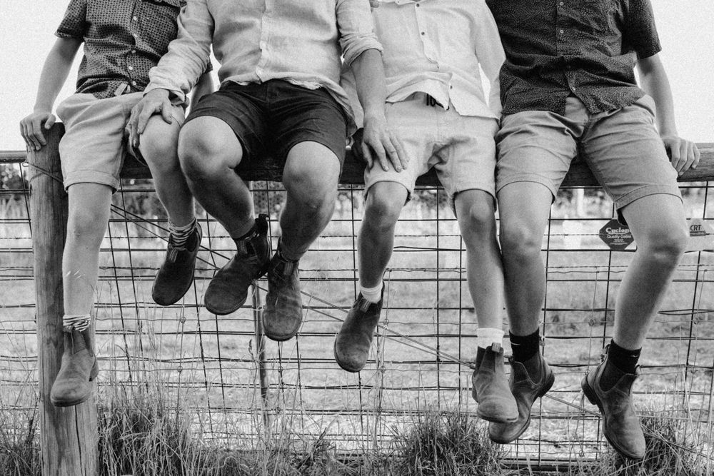 A black and white image of a farmer and his three sons boots - they are all sitting on a fence together for their family photoshoot.