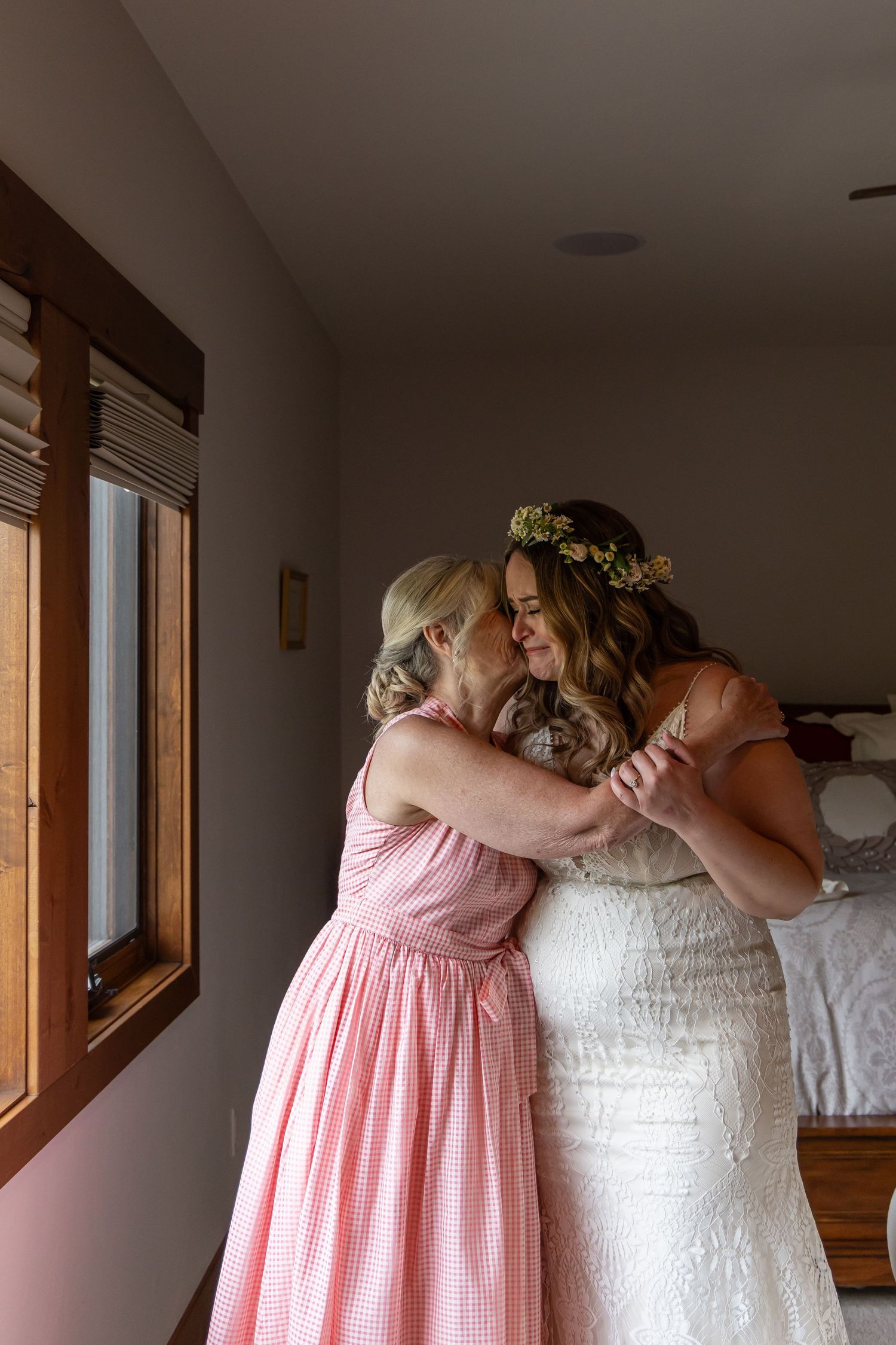 Emotional moment shared between a person in pink bridesmaid dress and bride in white gown embracing against natural window light.