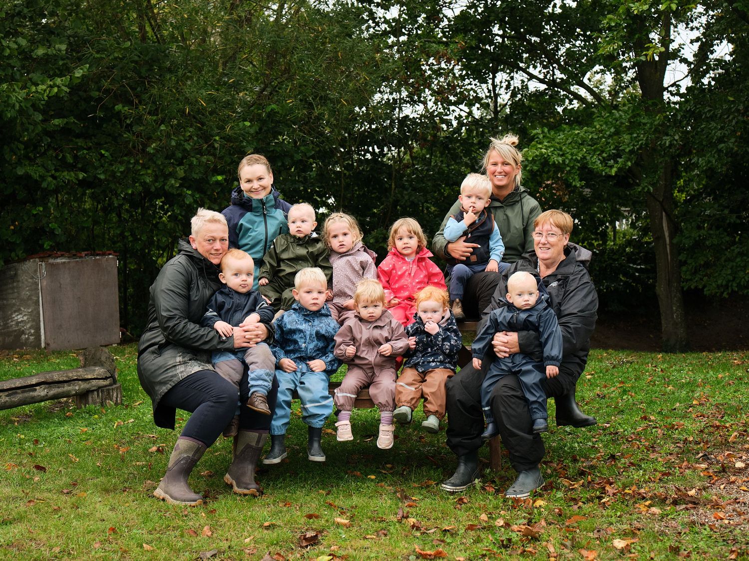 Group of adults and children sit together on wooden benches outdoors in an autumn forest setting.