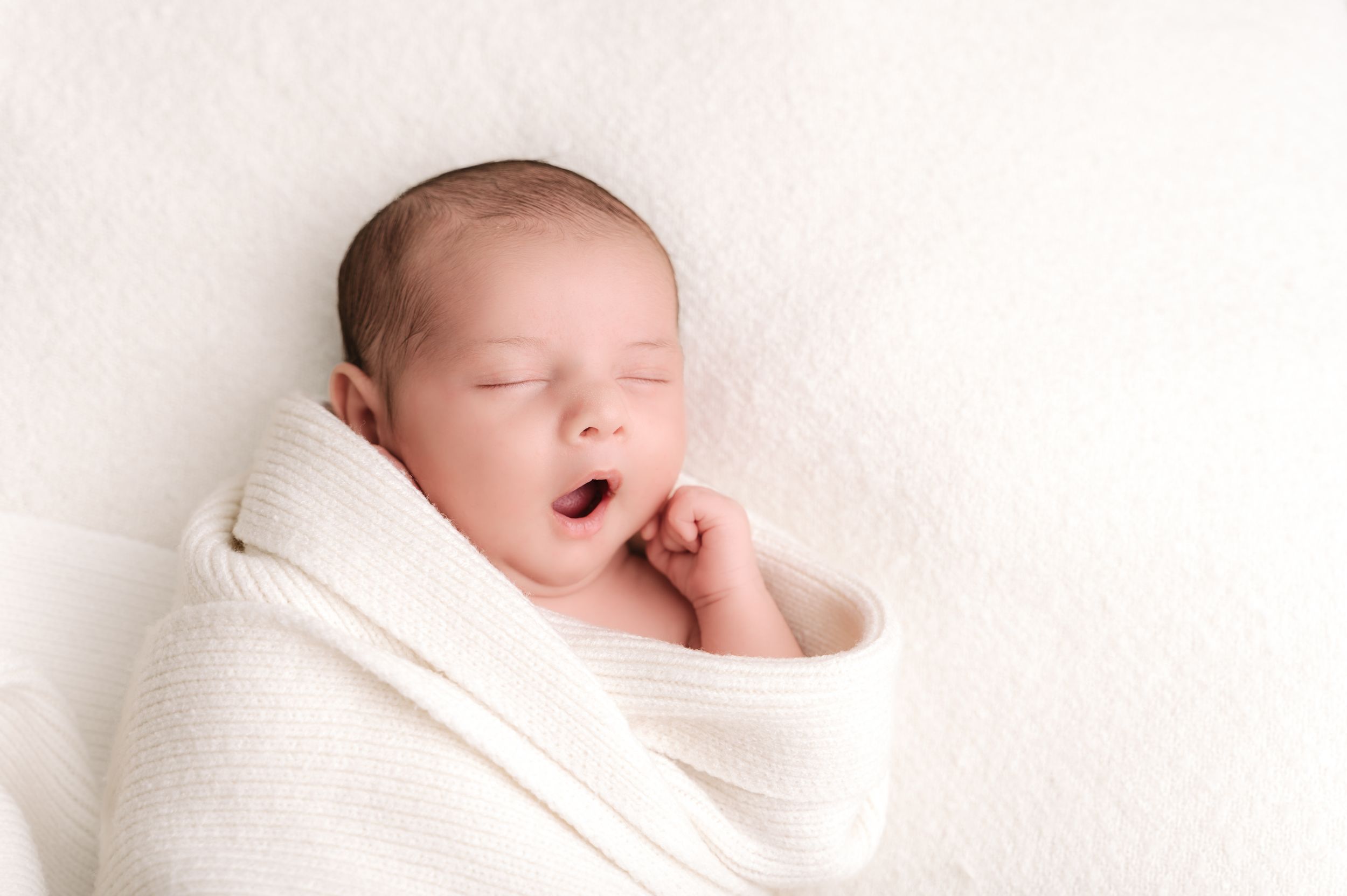 Peaceful newborn baby yawning while swaddled in a soft white blanket.