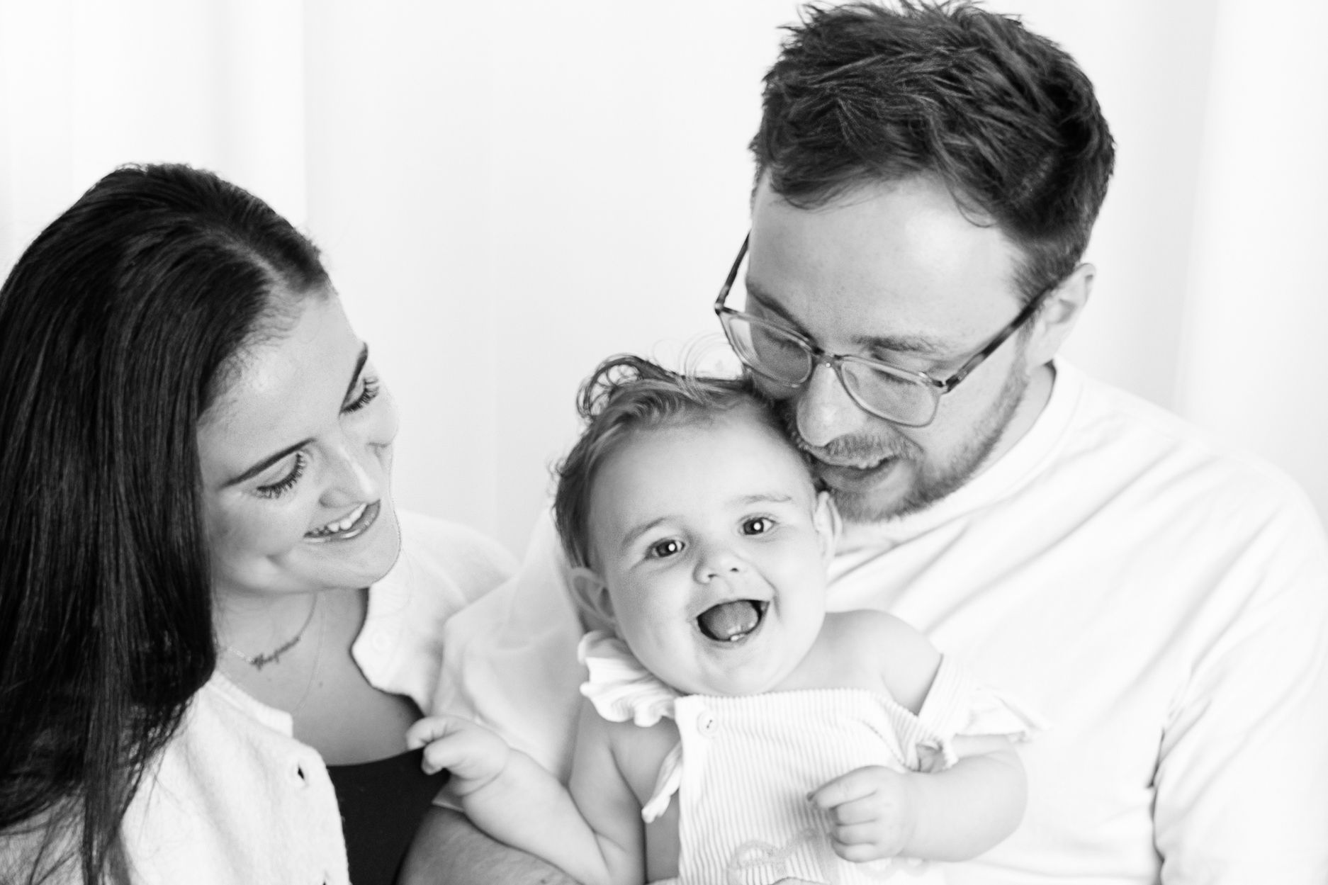 Black and white photo of smiling parents holding their laughing baby girl in a bright, cheerful family portrait.