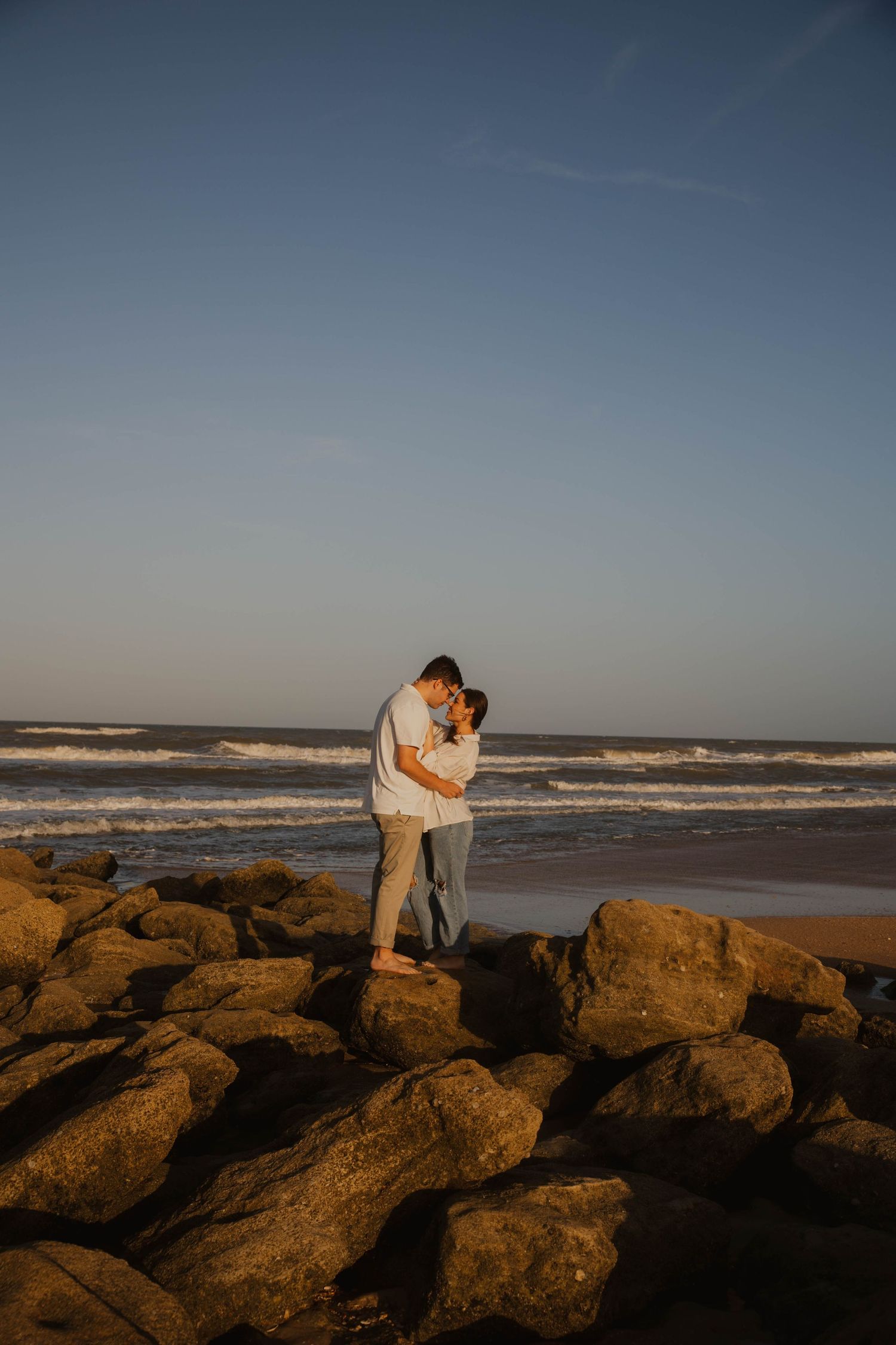 Sunset wedding photos of a couple embracing on coastal rocks with ocean waves in the background.