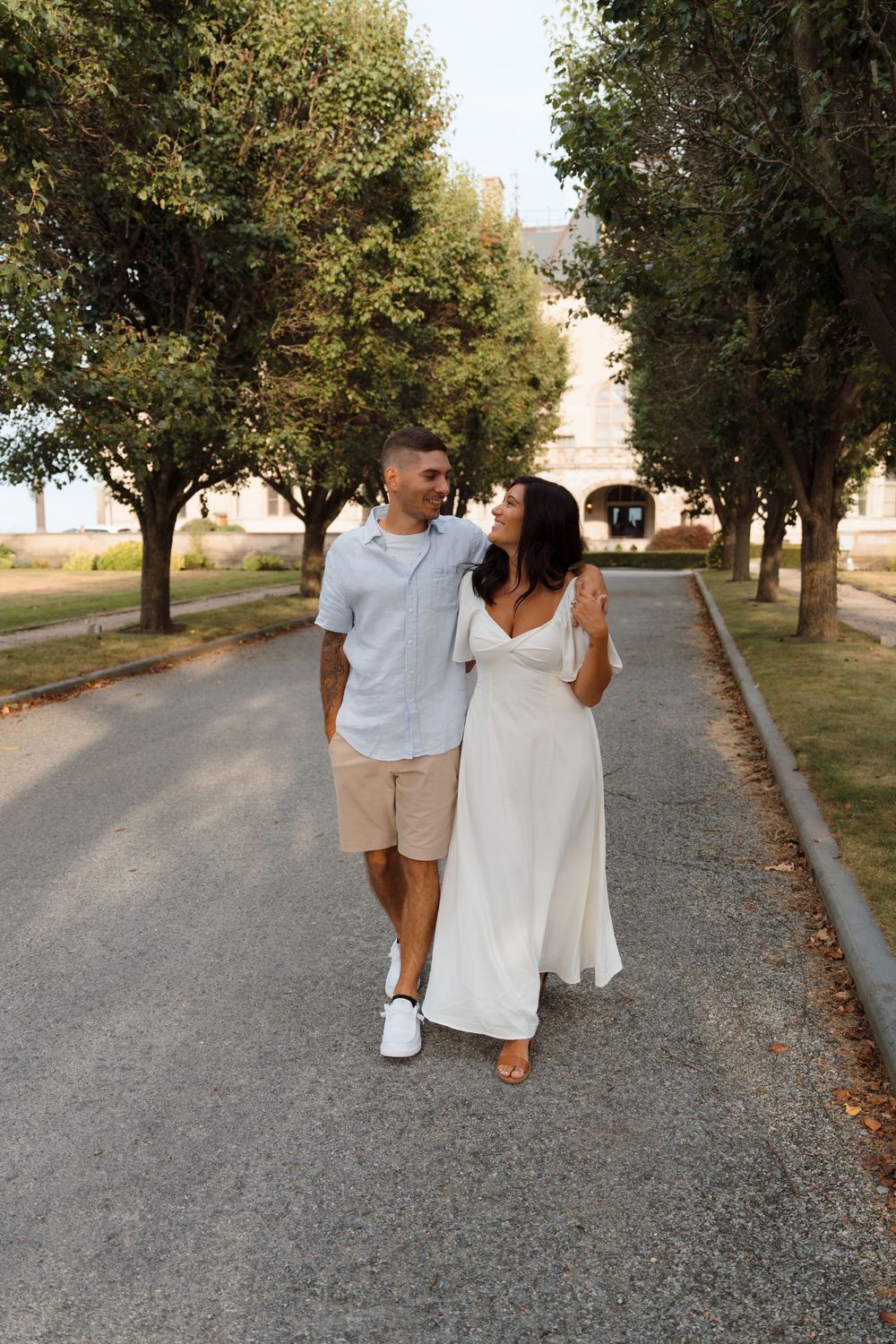 A couple walking together on a tree-lined gravel path wearing coordinated white and light blue outfits.