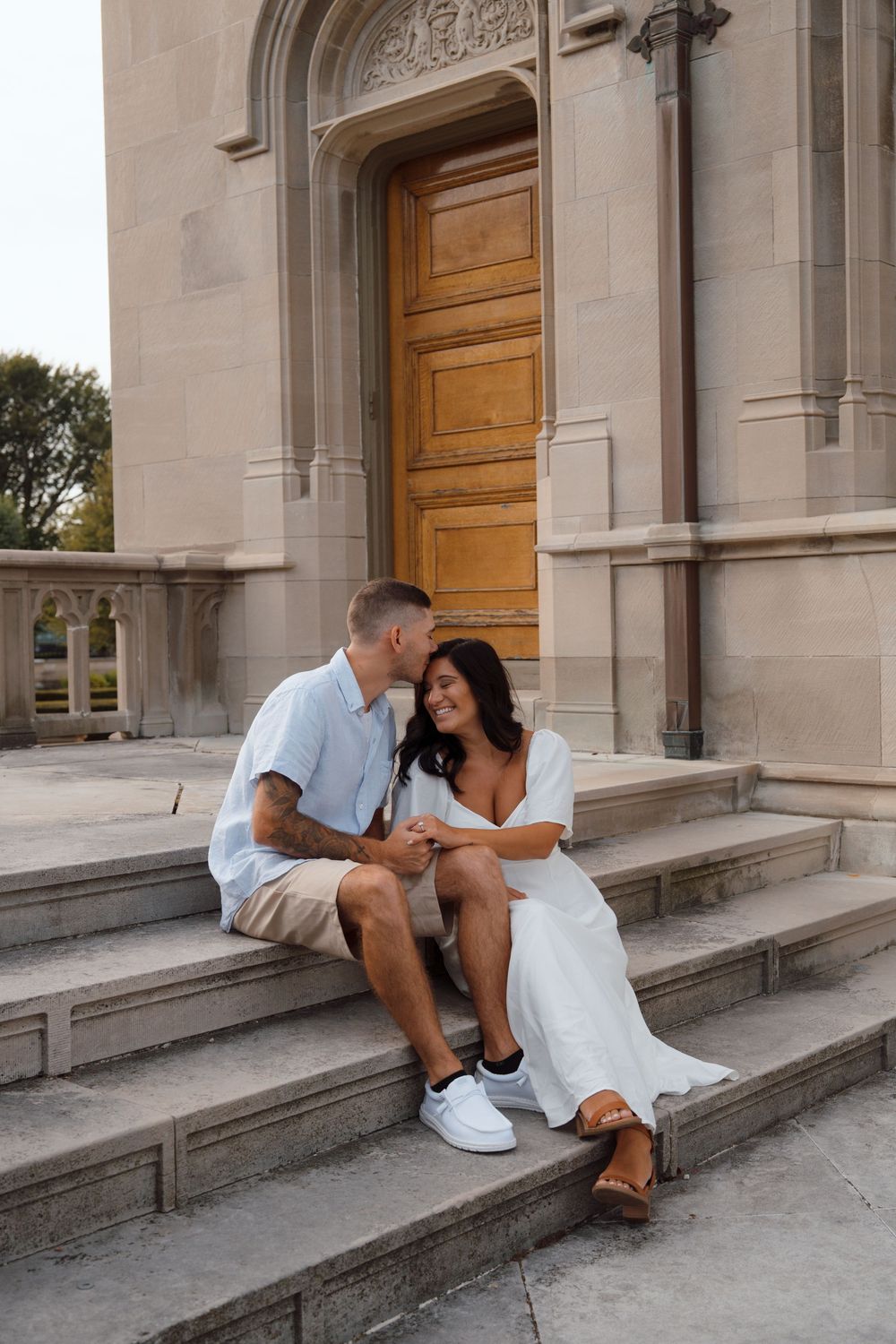 A couple shares an intimate moment sitting on stone steps in front of a grand wooden church door.