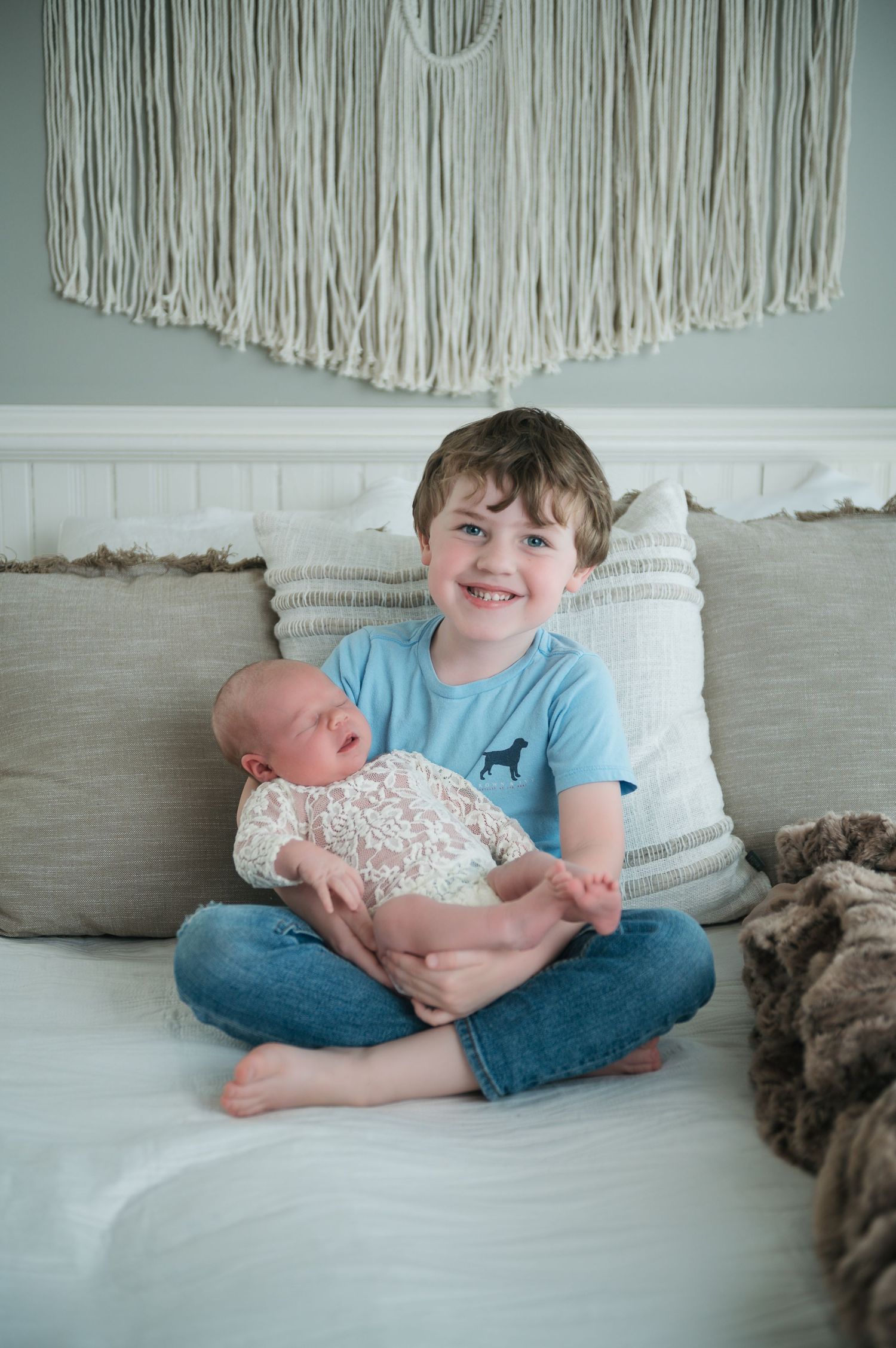 Children sitting on white bed with macrame wall hanging and cozy textiles in background.