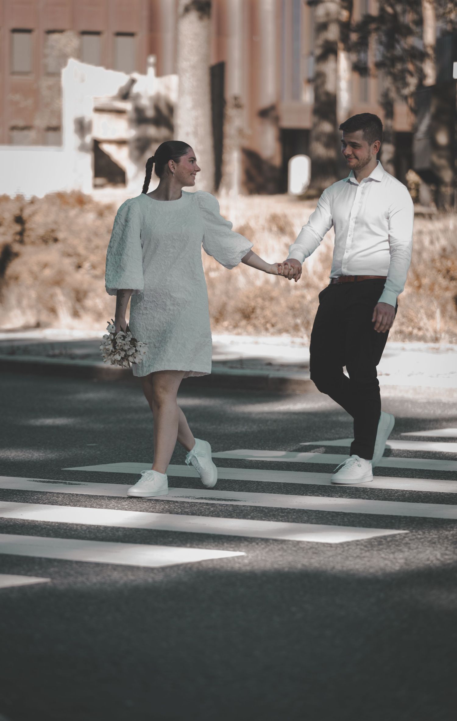 Bride and groom holding hands crossing a zebra crossing on their wedding day in matching white outfits.