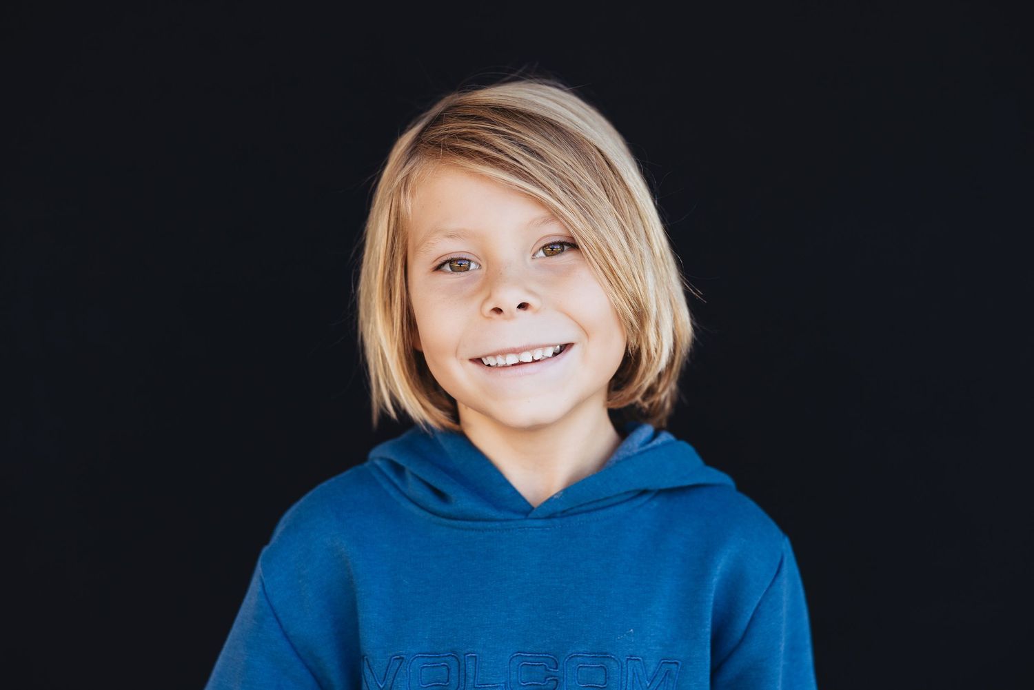 Portrait on dark background featuring a bright smile and blonde hair wearing a blue hoodie.