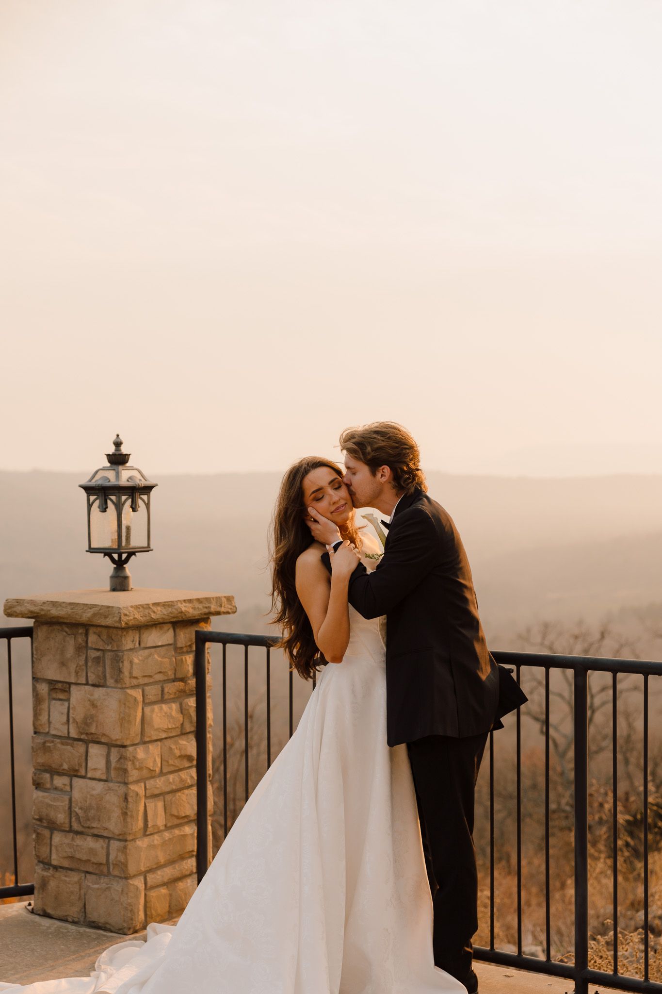 A bride and groom share a romantic moment on a stone balcony overlooking scenic hills at golden hour.