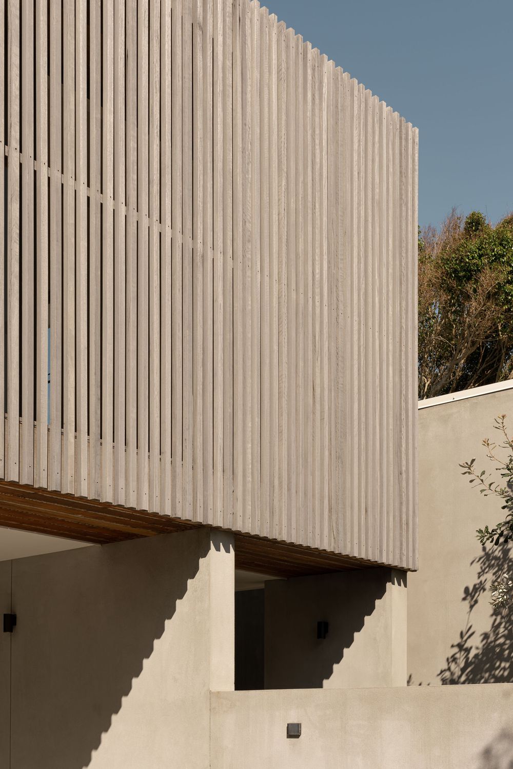 Modern architectural detail showing white vertical slats casting geometric shadows on a minimalist exterior wall.