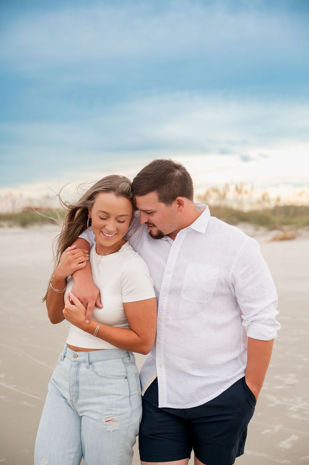 Joyful couple sharing laughter on sandy beach with dunes in background.