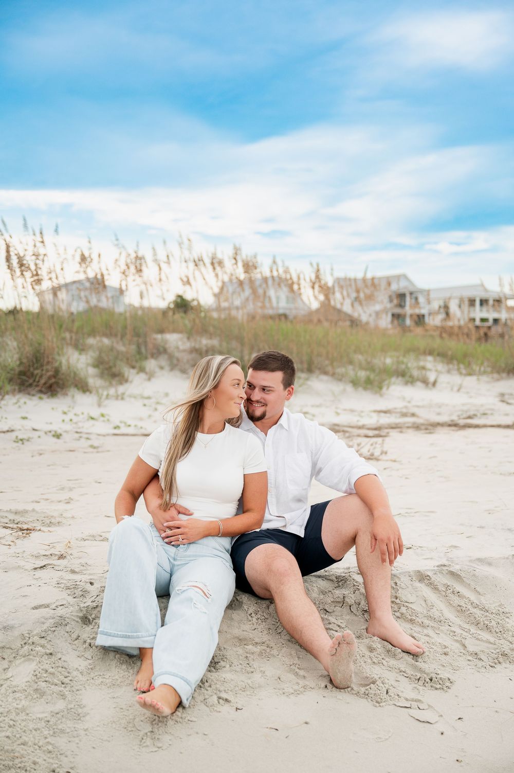 Couple relaxing on sandy beach with sea oats and coastal homes in background.