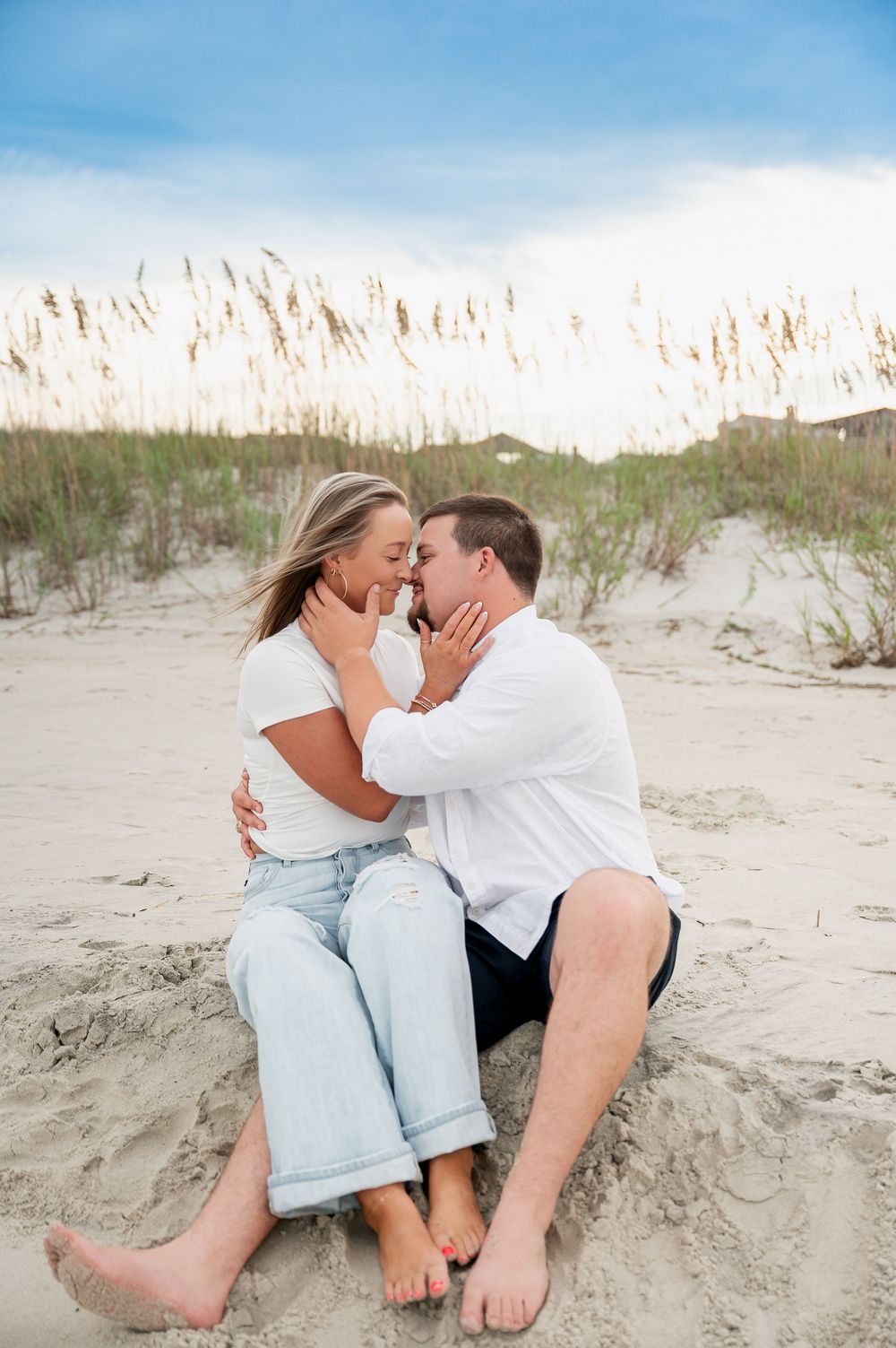 Intimate moment between couple on sandy beach with sea oats swaying in background.
