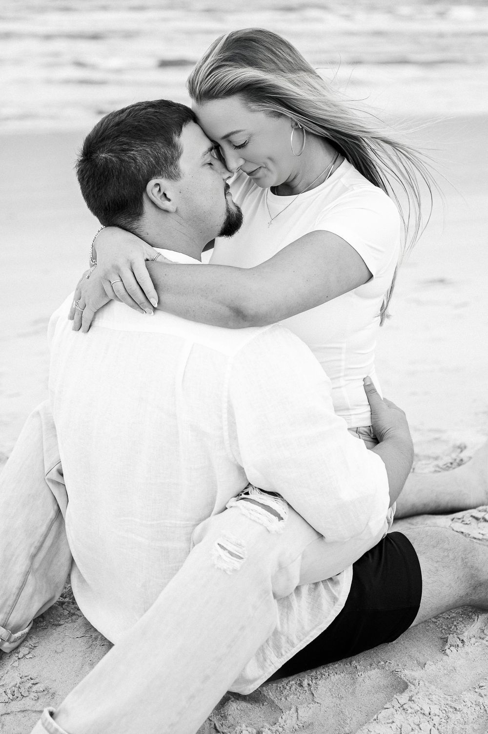 Romantic black and white beach photoshoot showing intimate couple poses in white attire on the sand.