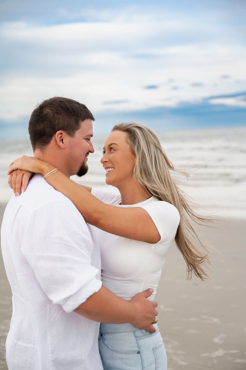 Couple embracing on beach with wind-swept hair and ocean backdrop.