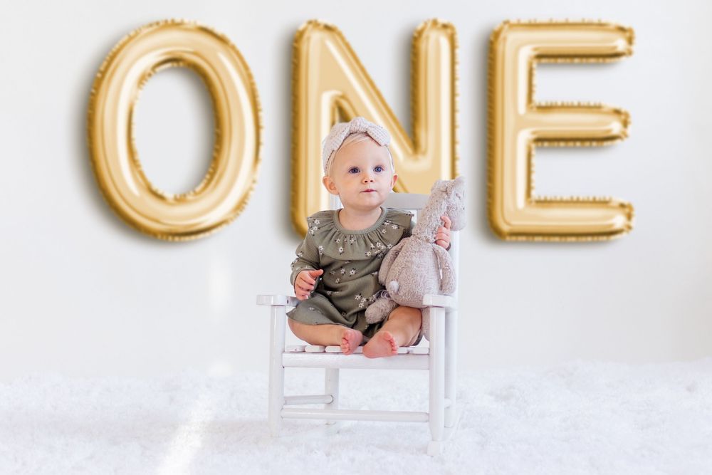 Gold metallic letter balloons spelling ONE with baby photos displayed against white wall for first birthday celebration.