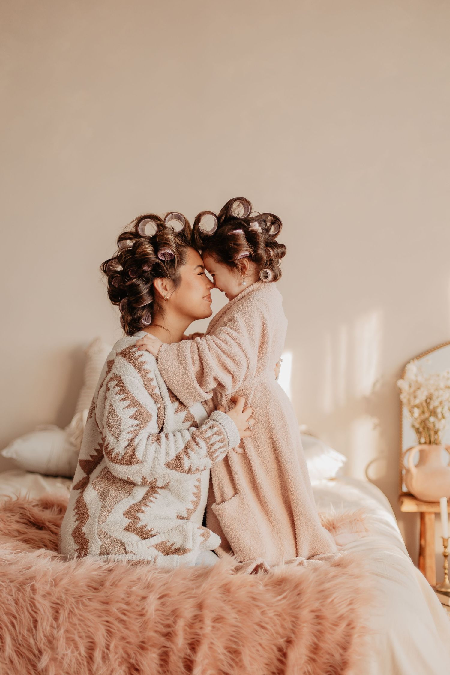 Mom and Daughter in curlers share a tender moment while sitting on a pink furry bed in soft lighting.