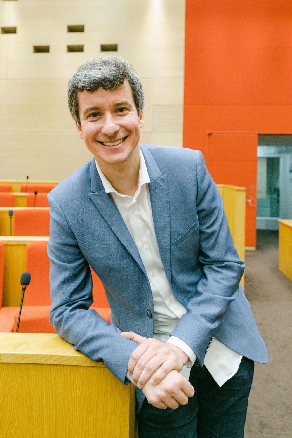 A person in a blue suit and white shirt sits at a table in a modern office setting with orange walls.