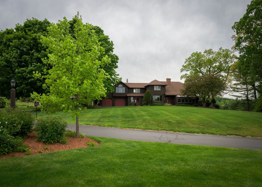 Large brick country estate home with manicured green lawn and mature trees under overcast spring sky.