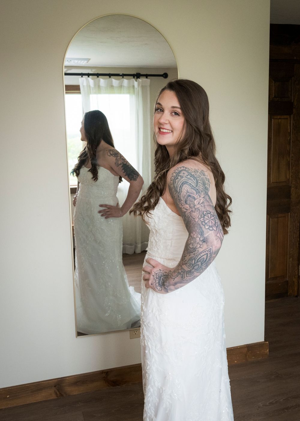 Tattooed bride in fitted lace wedding gown smiling at her reflection in a full-length arched mirror.