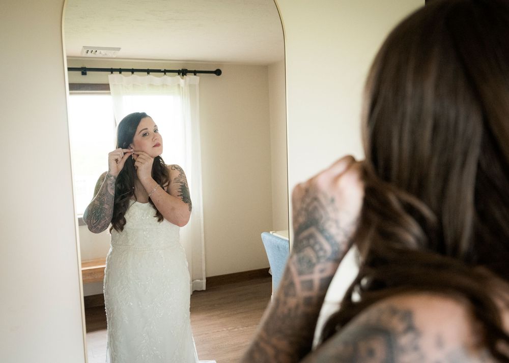 Tattooed bride adjusting her earring while looking in a full-length mirror while getting ready for her wedding.