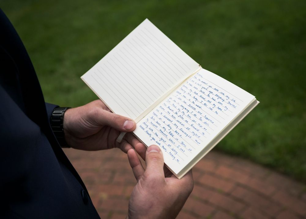 Groom reading handwritten vows from a small notebook outdoors on brick pavement.