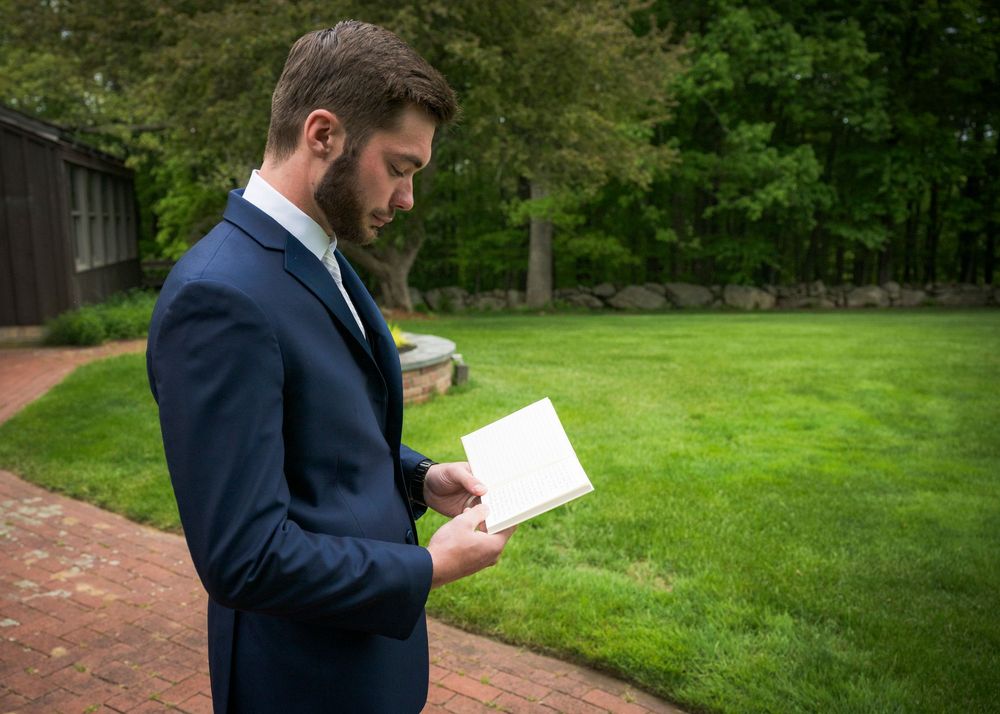 Groom in navy suit reading a card outdoors on a brick path surrounded by green lawn before wedding ceremony.