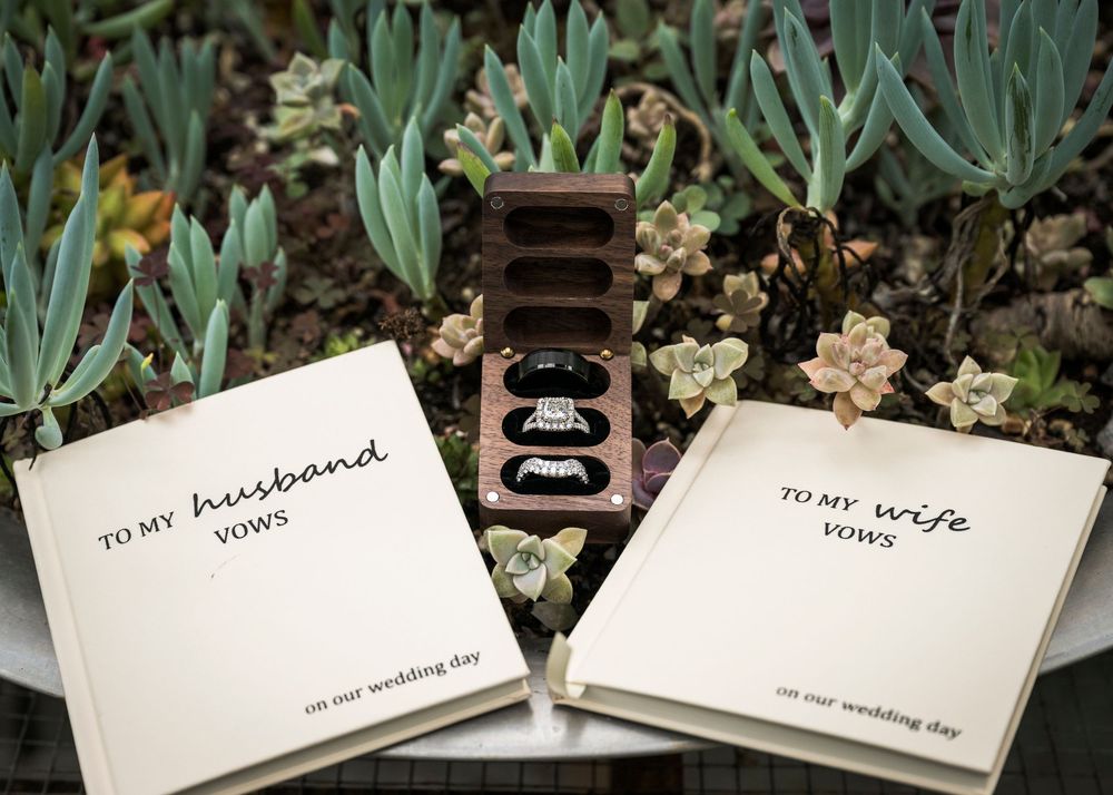 Wedding vow books labeled To My Husband and To My Wife displayed with wedding rings in a wooden box among succulents.