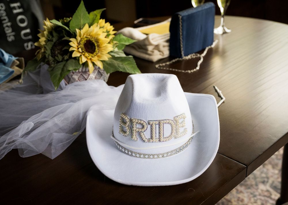 White cowboy hat with rhinestone BRIDE lettering on wooden table beside sunflowers and bridal veil.