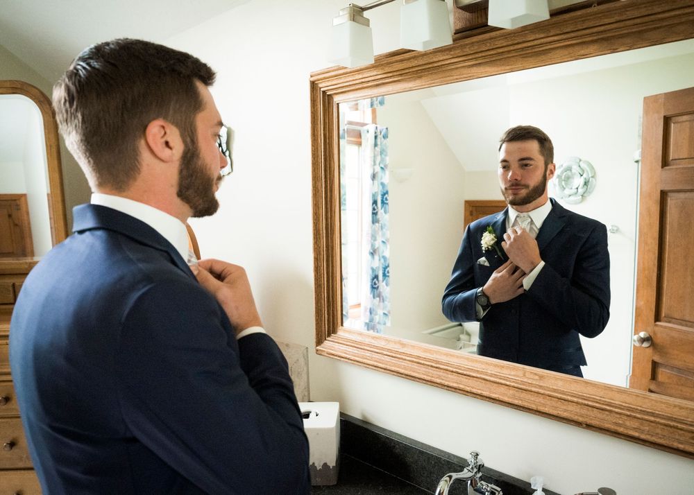 Groom in navy suit adjusting his tie while looking in a rustic wooden framed bathroom mirror before wedding.
