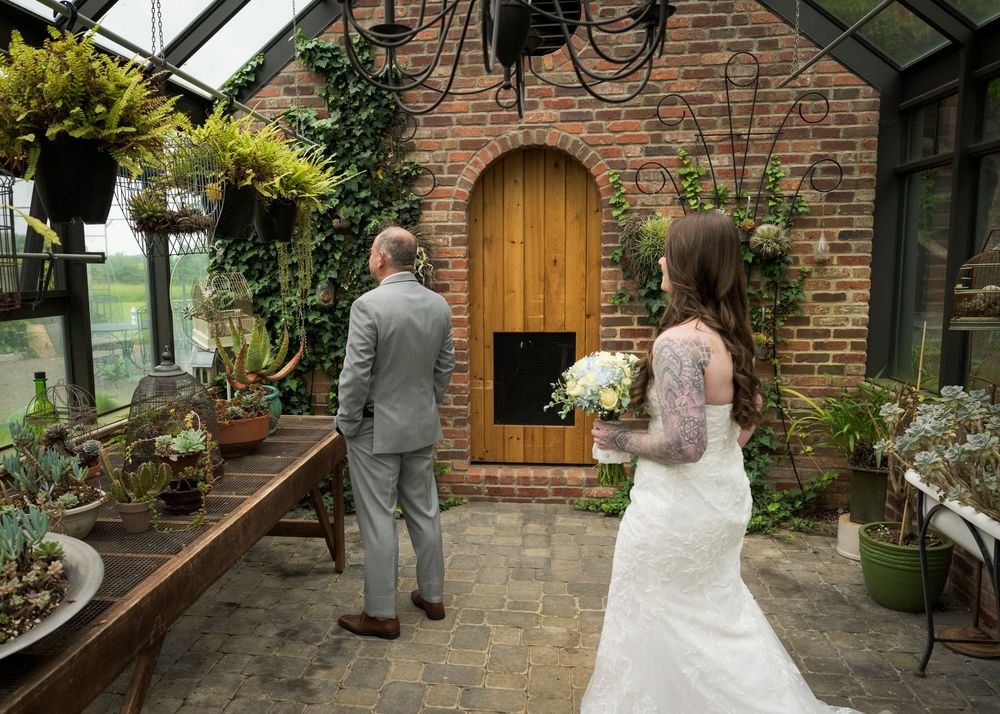 Bride in lace gown holding bouquet stands behind man in grey suit in rustic greenhouse venue.