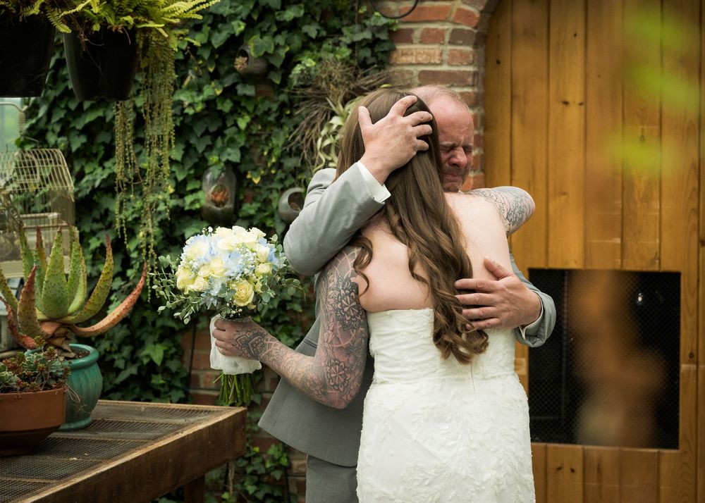 Emotional bride with floral tattoo embraces older man in gray suit outdoors near ivy-covered brick wall.