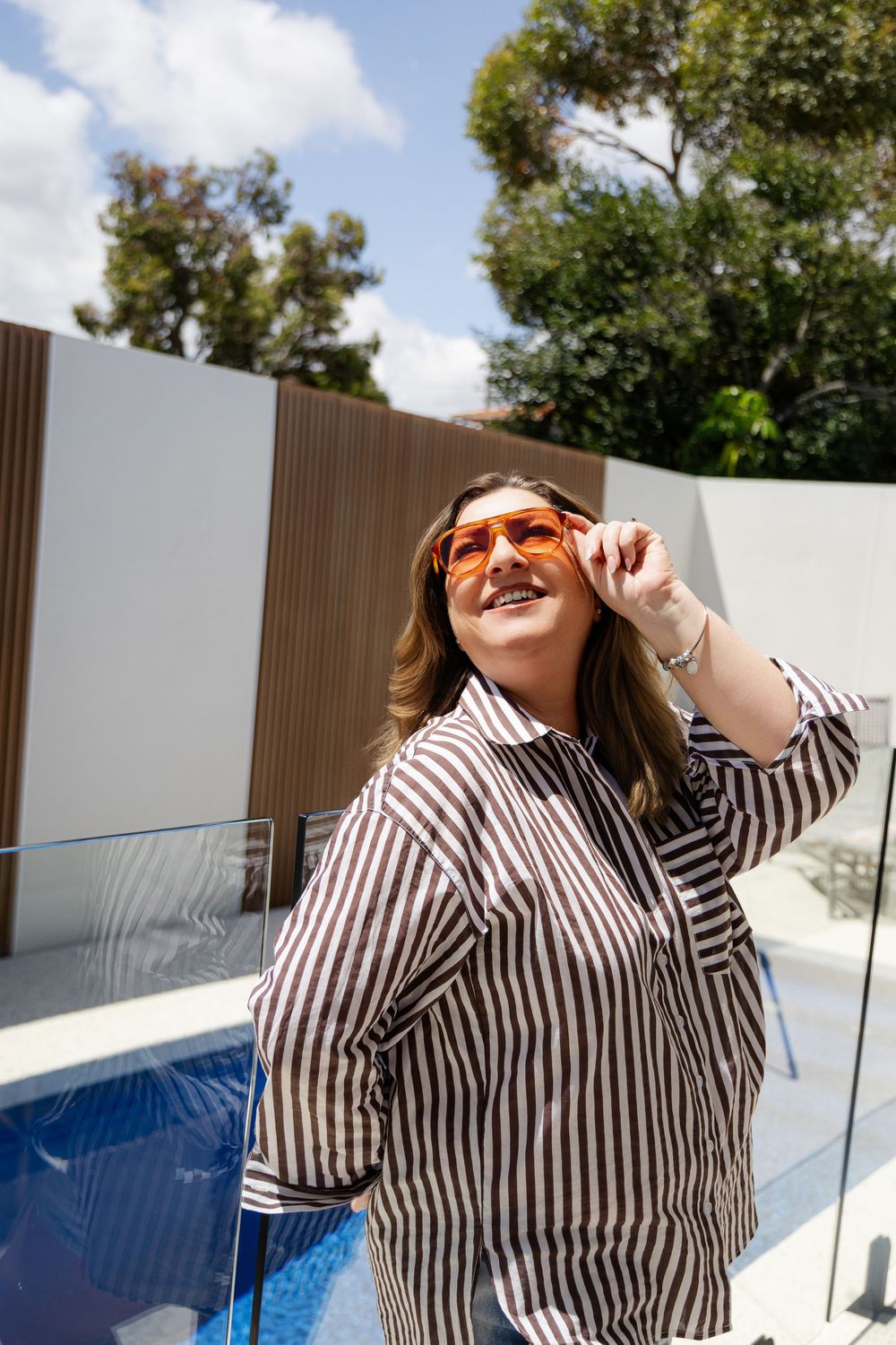 A person in a striped button-down shirt poses outdoors near a modern glass wall and swimming pool on a sunny day.