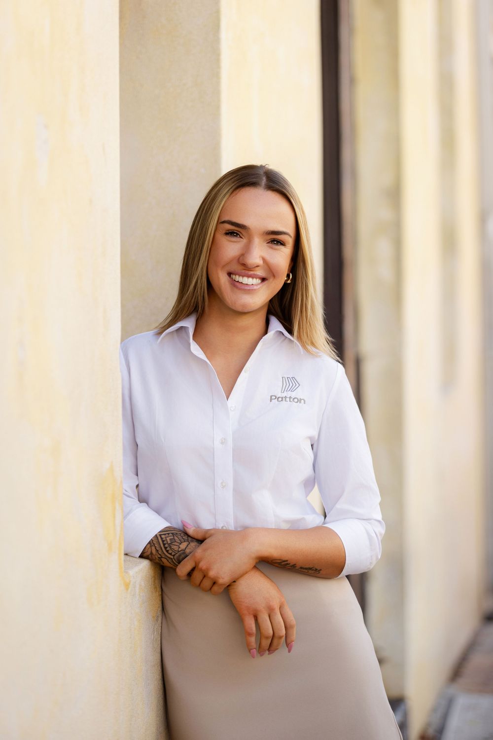 Professional portrait of someone in a crisp white button-down shirt with company logo leaning against a beige wall.