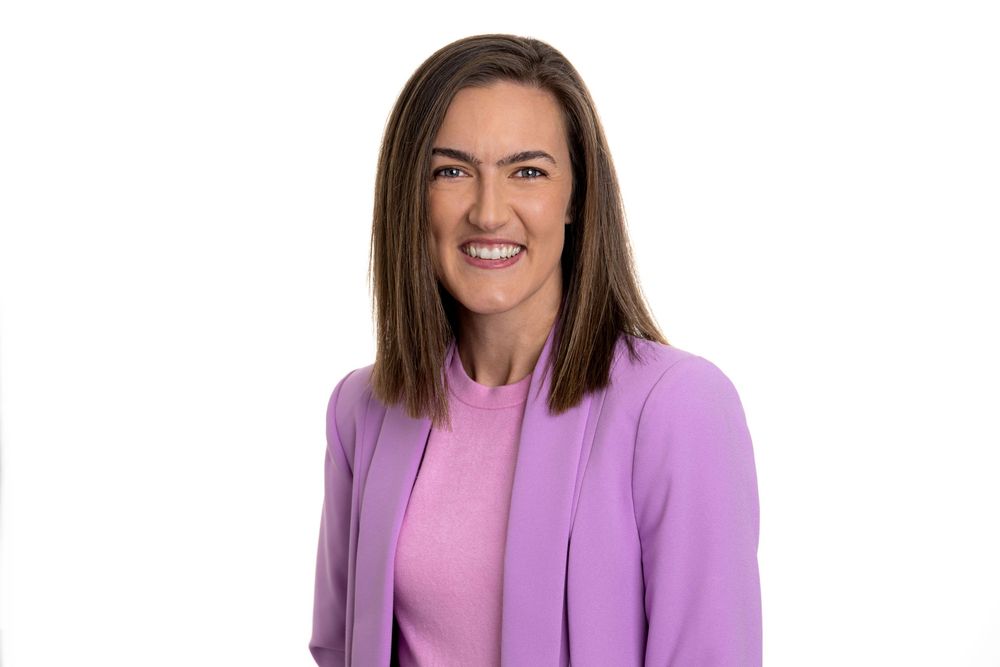 Professional wearing a vibrant purple blazer and pink top smiles warmly against a white background.