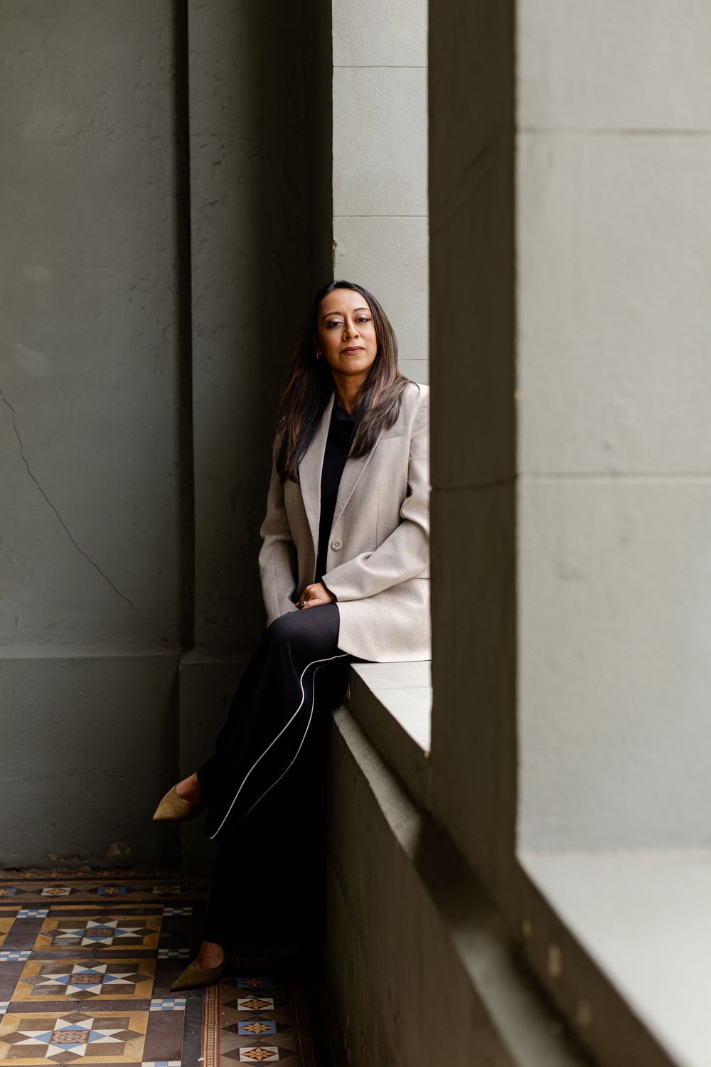 A person in a beige blazer sits thoughtfully by a window with patterned tile flooring in a moody architectural setting.