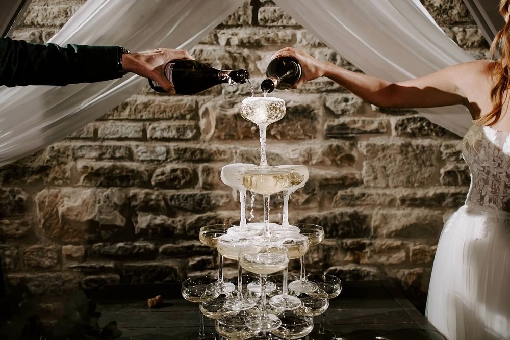Elegant champagne tower being filled at a wedding reception with crystal glasses catching dramatic lighting.