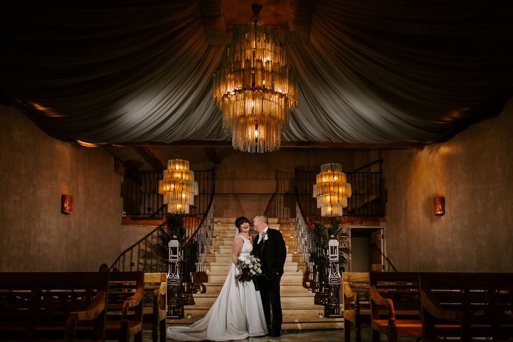 A couple stands on elegant stairs in a dimly lit venue with ornate chandeliers and dramatic draping.