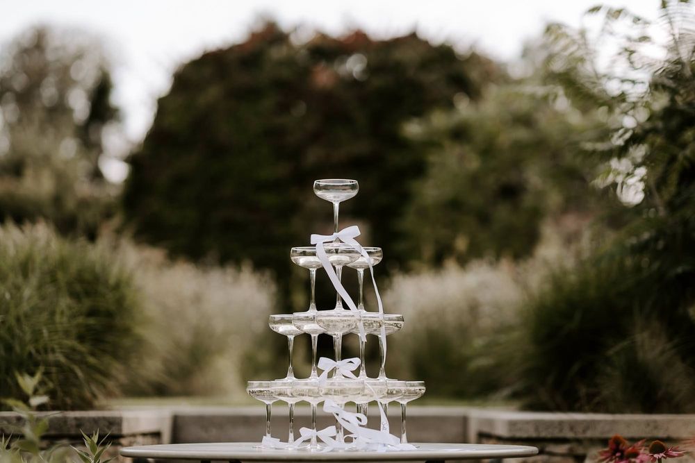 A champagne tower with glasses arranged in a pyramid shape stands elegantly outdoors at a wedding reception.