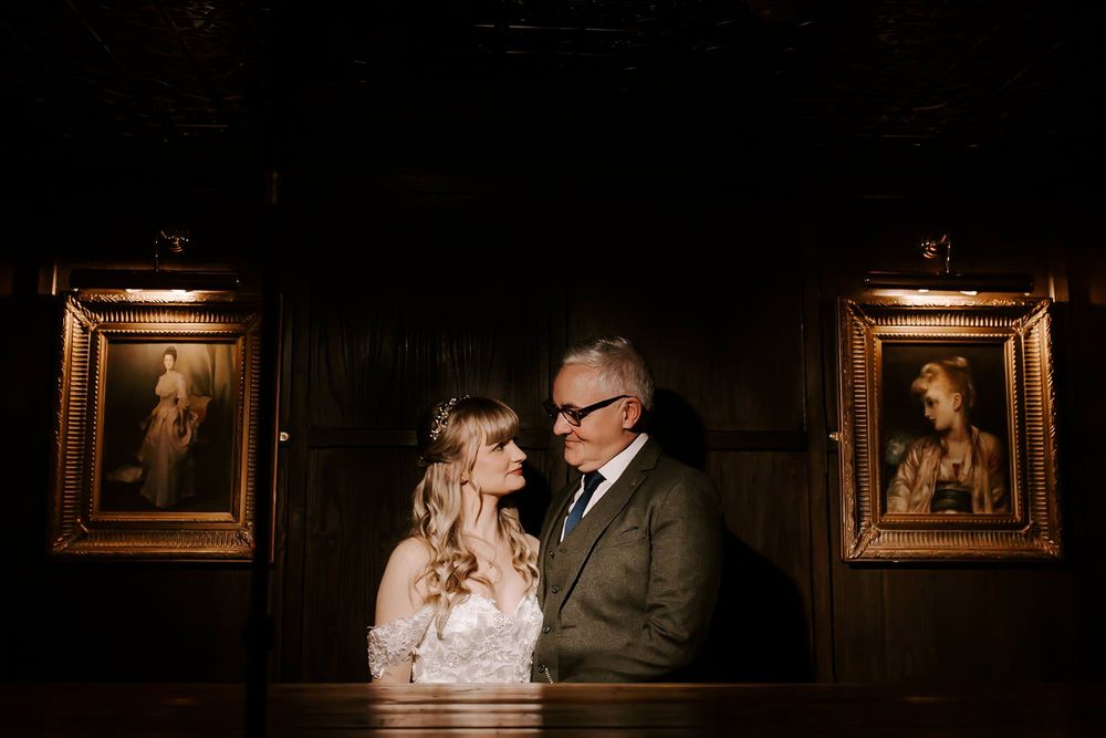 Romantic wedding couple poses in dimly lit art gallery with ornate gold frames on the walls.