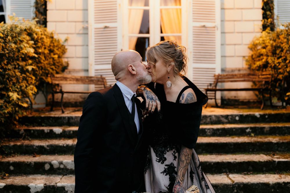 A couple embraces in front of a building with shutters during sunset with warm autumn lighting.
