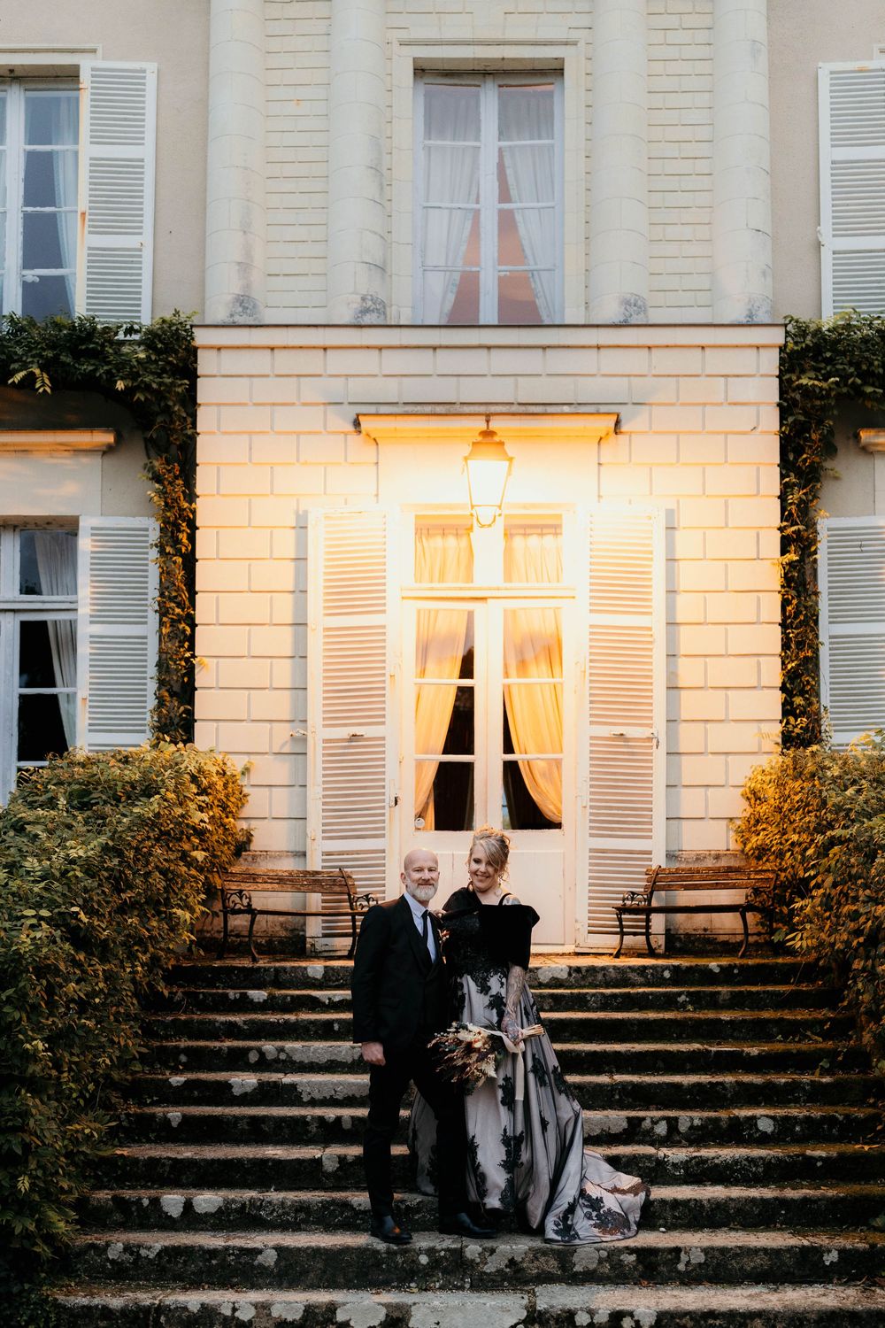 Elegant couple sits on stone steps outside a classic French chateau with white shuttered windows at sunset.