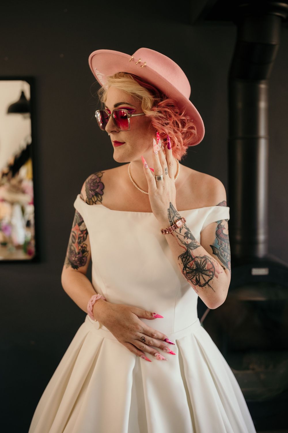 A person in a white wedding dress and pink hat shows off their arm tattoos while posing against a dark background.