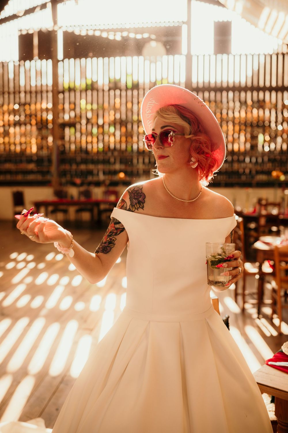 A vintage-inspired bride in an off-shoulder white dress and pink hat poses in golden sunlight streaming through windows.