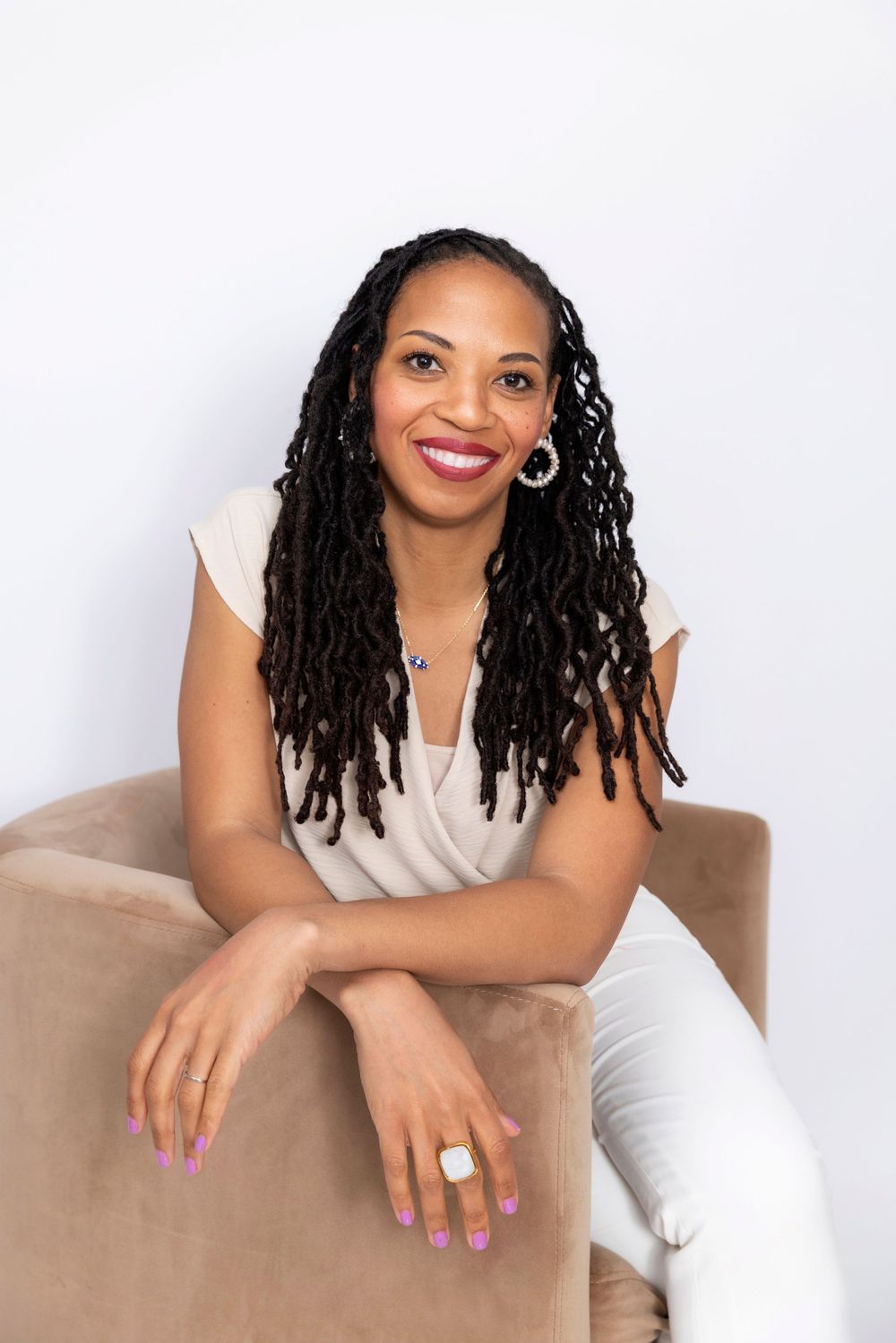 Professional portrait of someone with long locs wearing a white top and colorful nail polish seated on a beige armchair.
