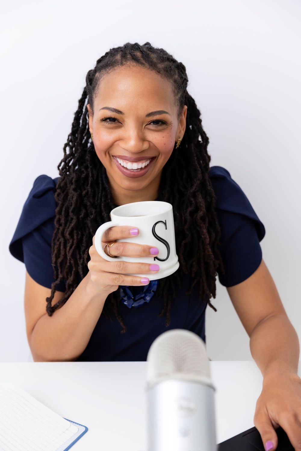 Woman with long locs wearing navy blue top holds white mug while sitting at desk with microphone and notebook.