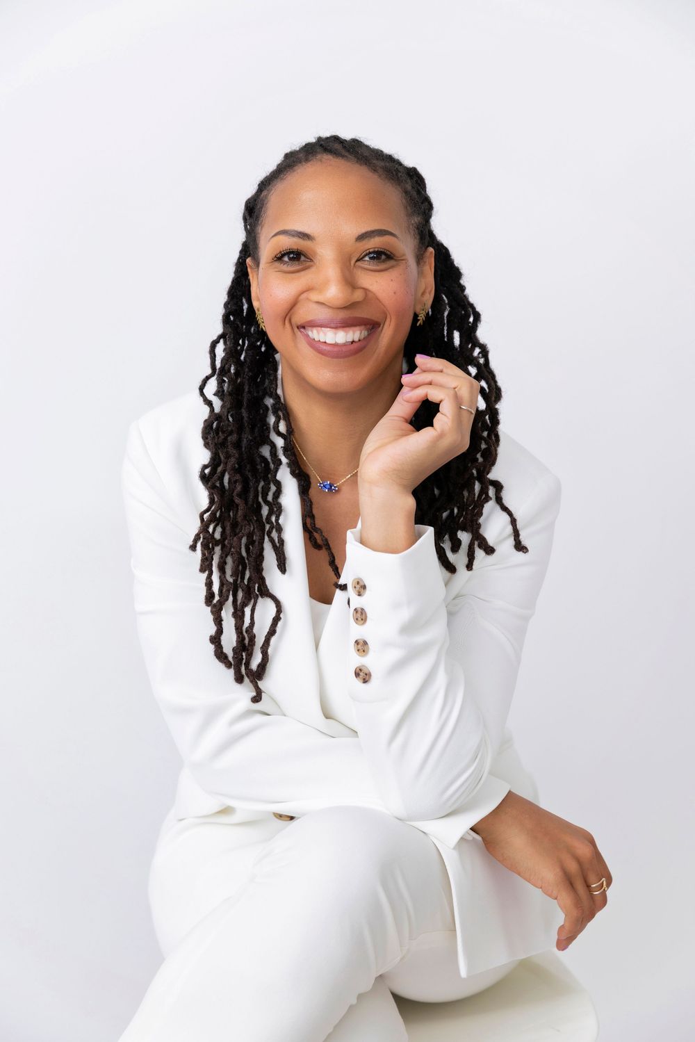 Professional studio portrait of a person with long locs wearing an all-white outfit against a light background.
