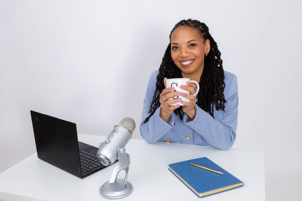 A content creator sits at a desk with podcast equipment, laptop and notebook while holding a coffee cup and smiling.